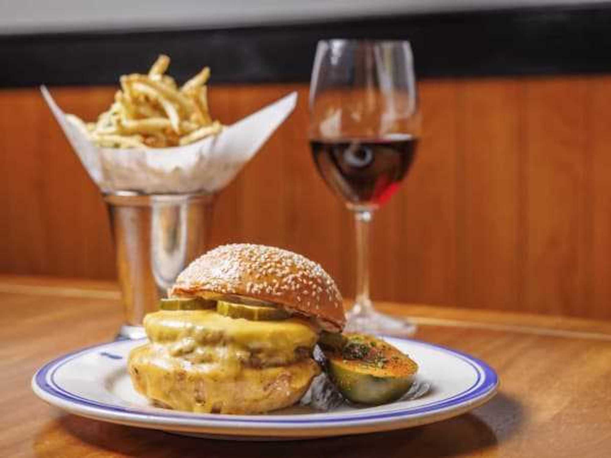 A cheeseburger on a white plate with fries and a glass of wine in the background